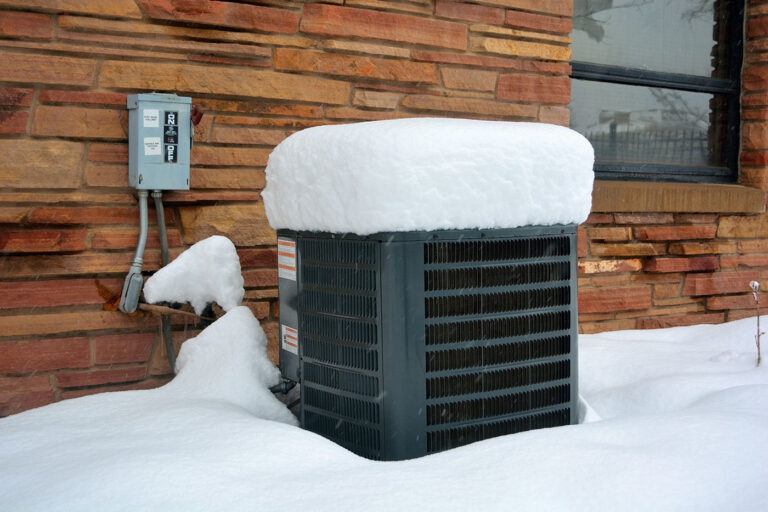 AC condenser covered in snow beside its outdoor disconnect box in winter.